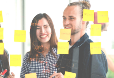  Diverse group of professionals in front of large, transparent presentation board covered with sticky notes
